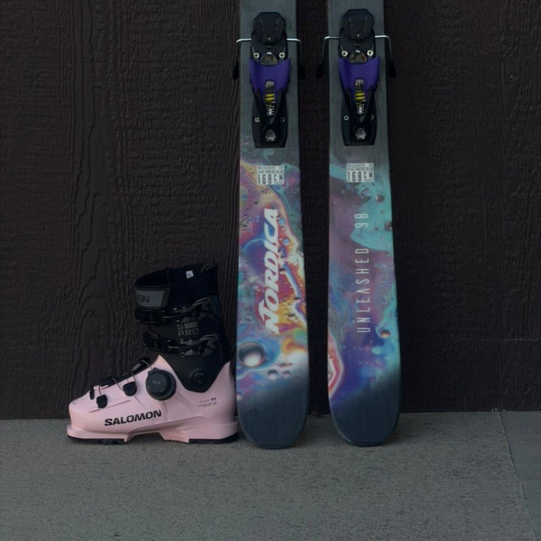 Two colorful skis with bindings and a pink Salomon ski boot against a dark background.